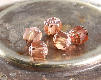 May include: Five rose gold colored glass beads with a faceted design. The beads are on a metallic surface.