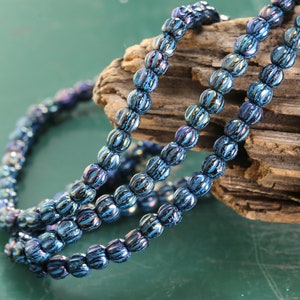 May include: Strands of iridescent blue beads with a ribbed texture. The beads are arranged in a curved pattern against a wooden background, creating a shimmering effect. The beads are likely used for jewelry making.