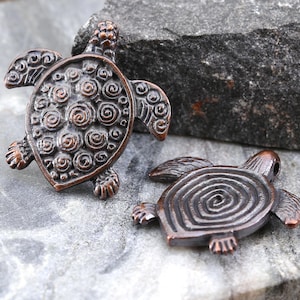May include: Two bronze-colored turtle pendants. One turtle has a detailed shell with spiral and swirl patterns. The other turtle has a shell with a spiral design. Both have small feet and heads. The pendants are on a gray stone surface.