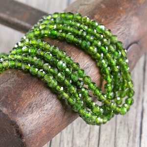 May include: A strand of faceted, bright green beads, likely gemstones, coiled on a piece of rough, dark wood. The beads have a sparkling, polished appearance, reflecting light. The overall effect is one of natural beauty and elegance.