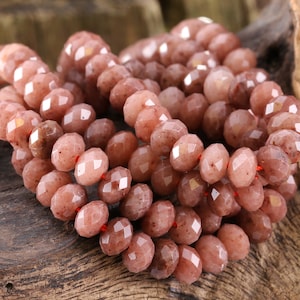 May include: A strand of faceted gemstone beads in shades of pink and brown. The beads are strung on a red thread and have a slightly irregular shape.