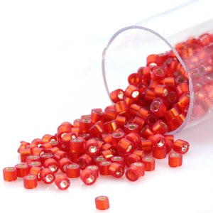 May include: A clear plastic tube filled with small, red, iridescent glass beads. The beads are spilling out of the tube onto a white surface.
