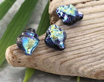 May include: Three iridescent, dark-colored seashell beads with a textured surface. The shells reflect a spectrum of colors, including blue, green, and yellow. They are arranged on a piece of driftwood, suggesting a coastal or nautical theme.