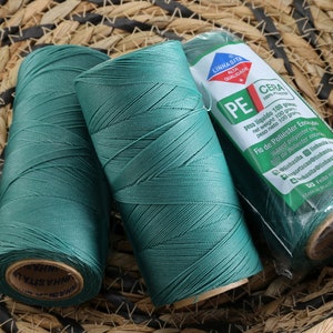 May include: Three spools of teal green waxed polyester thread. The spools are labeled "Linhasita Alta Qualidade PE/Cera 100gr"