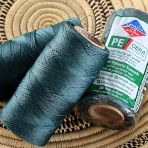 May include: Two spools of dark green waxed polyester thread, one is still in its packaging. The packaging is labeled "Linhasita Cera PE 100% Poliéster" and includes information about the product in Portuguese and Spanish. The spools are sitting on a woven basket.
