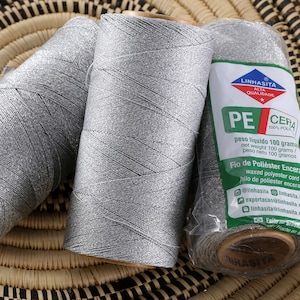 May include: Three spools of silver gray waxed polyester thread, each with a label that reads "Linhasita Alta Qualidade PE Cera 100% Poliester", "Peso Liquido 100 gramas", "Net Weight 100 grams", "Peso Neto 100 gramos", "Fio de Poliester Encerado", "Waxed Polyester Cord", "Hilo de Poliester Encerado", "@linhasita linhasita.com.br", "exportacao@linhasita.com.br", "linhasita@linhasita.com.br", "Feito no Brasil", "Hecho en Brasil", and "Linhasita".