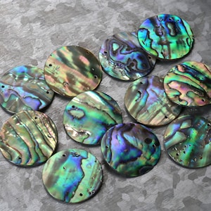 May include: A collection of iridescent abalone shell discs. The discs are round with a hole drilled near the edge. The shells display a range of colors, including green, blue, purple, and gold, with a shimmering, pearlescent effect. These are suitable for jewelry making.
