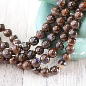 May include: Strands of brown, round beads with a glossy finish. The beads have a mix of colors, including brown, blue, and cream. The beads are resting on a light-colored wooden surface.