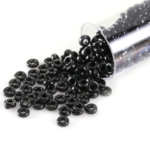 May include: A collection of small, black, donut-shaped beads spilling from a clear plastic tube. The beads are glossy and evenly sized, creating a visually appealing contrast against the white background. Ideal for jewelry making and crafting.