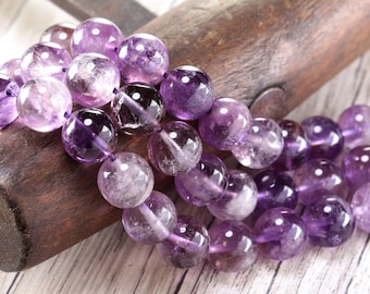 May include: Close-up of a strand of amethyst gemstone beads. The beads are round, translucent, and range in color from light lavender to deep purple. They are strung together on a purple cord, and rest on a wooden surface.