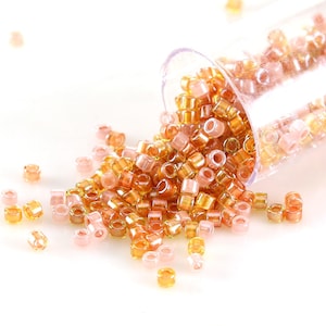 May include: A collection of small, cylindrical beads in shades of orange, gold, and pink, spilling from a clear plastic tube. The beads are scattered on a white surface, creating a visually appealing display for crafting or jewelry making.