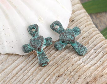 May include: Two rustic, turquoise-colored cross pendants with a heart-shaped center. The crosses have a weathered, antique appearance, with a loop at the top for hanging. The pendants are displayed on a light-colored, textured surface.