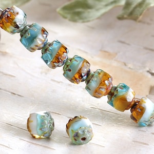 May include: Close-up of a strand of artisan glass beads. The beads are a mix of white, teal, and amber colors. Each bead has a faceted, barrel shape. The beads are strung on a silver-colored wire, perfect for jewelry making.