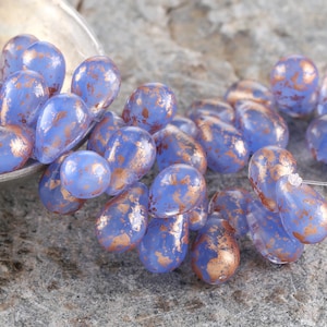 Peut inclure: Un ensemble de perles bleues en forme de goutte d'eau avec un motif tacheté de couleur cuivre métallique. Les perles sont dispersées sur une surface de pierre grise, certaines sont dans une cuillère argentée. Idéal pour la création de bijoux.