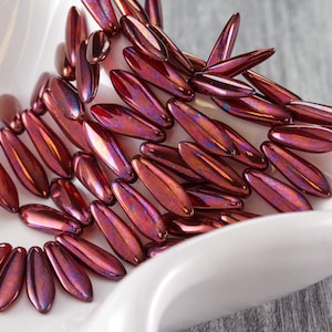 May include: A pile of iridescent burgundy glass beads in a leaf shape. The beads are arranged in a fan shape on a white surface.