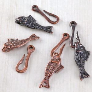 May include: Close-up of several metal fish-shaped clasps and hook closures. The clasps are in shades of copper and dark gray, with detailed fish scales and fins. The hooks are curved and attached to the fish. These are likely used for jewelry or craft projects.