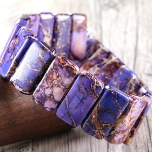 May include: A stretch bracelet featuring rectangular, polished stones in shades of purple, lavender, and brown. The bracelet is made of natural stone and has a marbled appearance. The stones are strung together to form a flexible band.