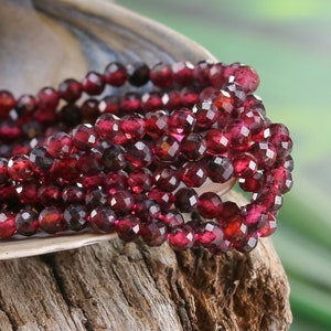 May include: A strand of deep red garnet beads, faceted and polished, arranged in a coil on a wooden surface. The beads are small and uniform in size, creating a rich and elegant look.