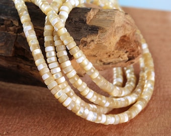 Peut inclure: Un brin de perles de coquillage blanches et beiges, avec une finition légèrement irisée. Les perles sont rondes et ont une forme légèrement irrégulière. Les perles sont enfilées sur un fil transparent et reposent sur un morceau de bois.