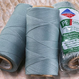 May include: Three spools of light blue waxed polyester cord, each wrapped in brown paper with a label that reads "Linhasita Alta Qualidade PE Cera 100% Poliéster" and "Fio de Poliéster Encerado" in Portuguese. The label also includes the text "Feito no Brasil" and "Made in Brazil / Hecho en Brasil".