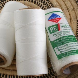 May include: Three spools of white waxed polyester cord, each with a label that reads "Linhasita PE Cera 100% Poliester" and includes information about the product in Portuguese and Spanish. The label also includes the text "Feito no Brasil" which means "Made in Brazil".