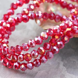 May include: Close-up of strands of faceted, ruby-red beads. The beads have an iridescent quality, reflecting light with red, orange, and gold hues. The beads are strung together, suitable for jewellery making or crafts.