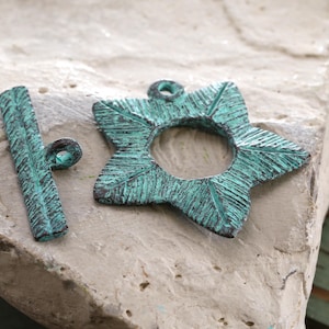 May include: Two antique-style metal jewelry components. One is a star-shaped pendant with a central hole, and the other is a bar with a loop. Both have a distressed teal finish with dark accents, suggesting an aged appearance.