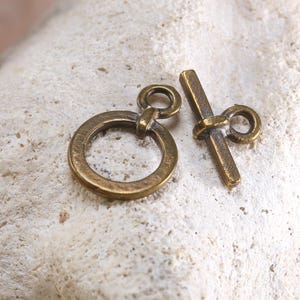 May include: A set of antique brass toggle clasps, featuring a circular ring and a bar with a loop. The clasps are designed for jewelry making and crafting. The metal has a warm, aged patina, suggesting vintage or handmade items.