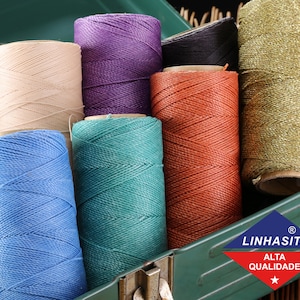 May include: A collection of spools of thread in various colors, including beige, purple, black, orange, blue, and green. The spools are arranged in a metal box with a green lid. The lid has a logo that says "Linhasita Alta Qualidade" with a star.