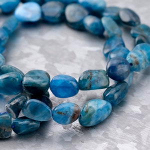 May include: A strand of blue apatite gemstone beads, tumbled and polished into smooth, oval shapes. The beads are strung together on a clear thread and are arranged in a loose, flowing pattern.