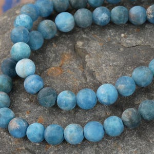 May include: A strand of blue agate beads, with a frosted, matte finish. The beads are round and uniform in size, and they are strung on a clear thread.