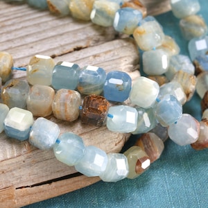 May include: Strands of faceted aquamarine beads in shades of blue, tan, and brown. The beads are strung on a blue cord and have a geometric, cube-like shape. The beads are displayed on a wooden surface.
