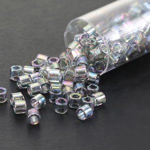 May include: A clear plastic tube filled with iridescent silver seed beads. The tube is tipped over and the beads are spilling out onto a black surface.
