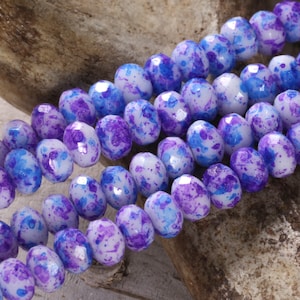 Peut inclure: Un brin de perles de verre blanches avec des éclaboussures de peinture bleue et violette. Les perles sont facettées et ont une finition brillante.