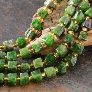 May include: Close-up of a necklace featuring vibrant green square-cut beads interspersed with small gold-colored beads. The necklace is displayed against a textured, dark background, highlighting the gemstone's color and shape. The beads are strung together, creating a layered effect.