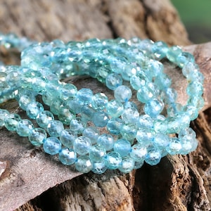 May include: A strand of light blue faceted gemstone beads. The beads are round and have a smooth, polished finish. The beads are strung on a clear thread and are arranged in a loose, flowing pattern.