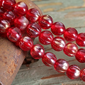 May include: Close-up of strands of red glass beads. Each bead is round with a ribbed design and a glossy finish. The beads are a deep, transparent red, catching the light and reflecting a warm glow. The background is a rustic, weathered surface.