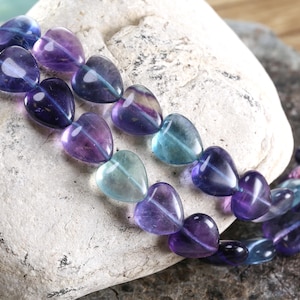 May include: A strand of heart-shaped beads in shades of purple, blue, and green. The beads are translucent and appear to be made of polished stone. The beads are resting on a light-colored, textured rock.