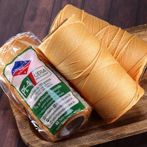 May include: Three spools of waxed polyester cord in a warm, golden-yellow hue. One spool is in a clear plastic wrapper with a green and white label that reads "LINHASITA". The spools are on a wooden tray with a dark brown wood grain.