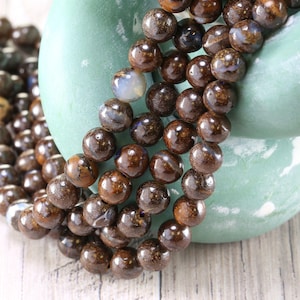 May include: Strands of brown gemstone beads with a speckled appearance. The beads are round and have a polished finish. The beads are displayed on a light green, rounded object, with a wooden surface in the background.