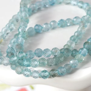 May include: A strand of light blue aquamarine beads. The beads are faceted and have a round shape. The beads are strung on a clear thread.