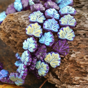 May include: Close-up of a strand of iridescent leaf-shaped beads. The beads have a gradient of colors, including purple, blue, and yellow. The beads are arranged on a piece of weathered wood.