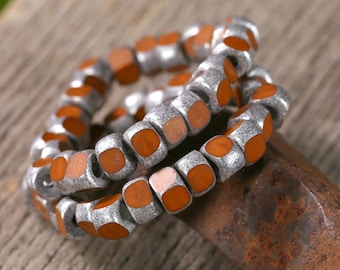 Puede incluir: Cadenas de cuentas plateadas con centros cuadrados de color naranja-marrón. Las cuentas están dispuestas en un patrón circular, creando un efecto de capas. Las cuentas tienen un aspecto texturizado y metálico, y los centros naranjas proporcionan contraste.
