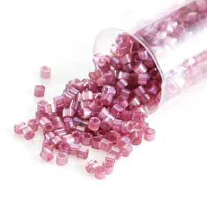 May include: A collection of small, cylindrical, pink beads spilling from a clear plastic tube. The beads have a subtle iridescent sheen, reflecting light. The beads are scattered on a white surface, suggesting a craft or jewelry-making theme.