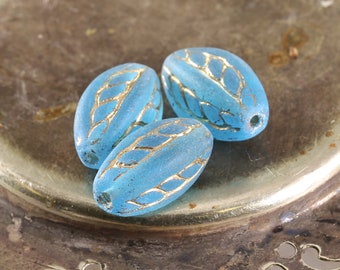 May include: Three oval-shaped glass beads with a light blue color and gold leaf designs. The beads are on a silver surface.