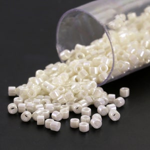 May include: A collection of small, off-white, cylindrical beads spilling from a clear plastic tube. The beads have a subtle iridescent sheen, suggesting a pearlescent finish. Ideal for jewelry making and crafting projects.