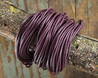 May include: A bundle of dark purple leather cord coiled on a weathered wooden branch. The cord appears smooth and round, with a slight sheen. The wood has a rough texture with patches of green and yellow lichen.
