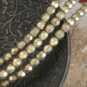 May include: A strand of gold-toned faceted beads, arranged in a bowl. The beads are round and have a metallic finish.