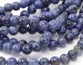 May include: Close-up of strands of round, blue sodalite gemstone beads. The beads have a mottled appearance with darker blue and black inclusions. The beads are strung together, ready for jewelry making or crafting projects.