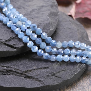 May include: Three strands of faceted, light blue gemstone beads. The beads are small, round, and have a sparkling, polished appearance. The strands are arranged on a dark gray, textured surface.